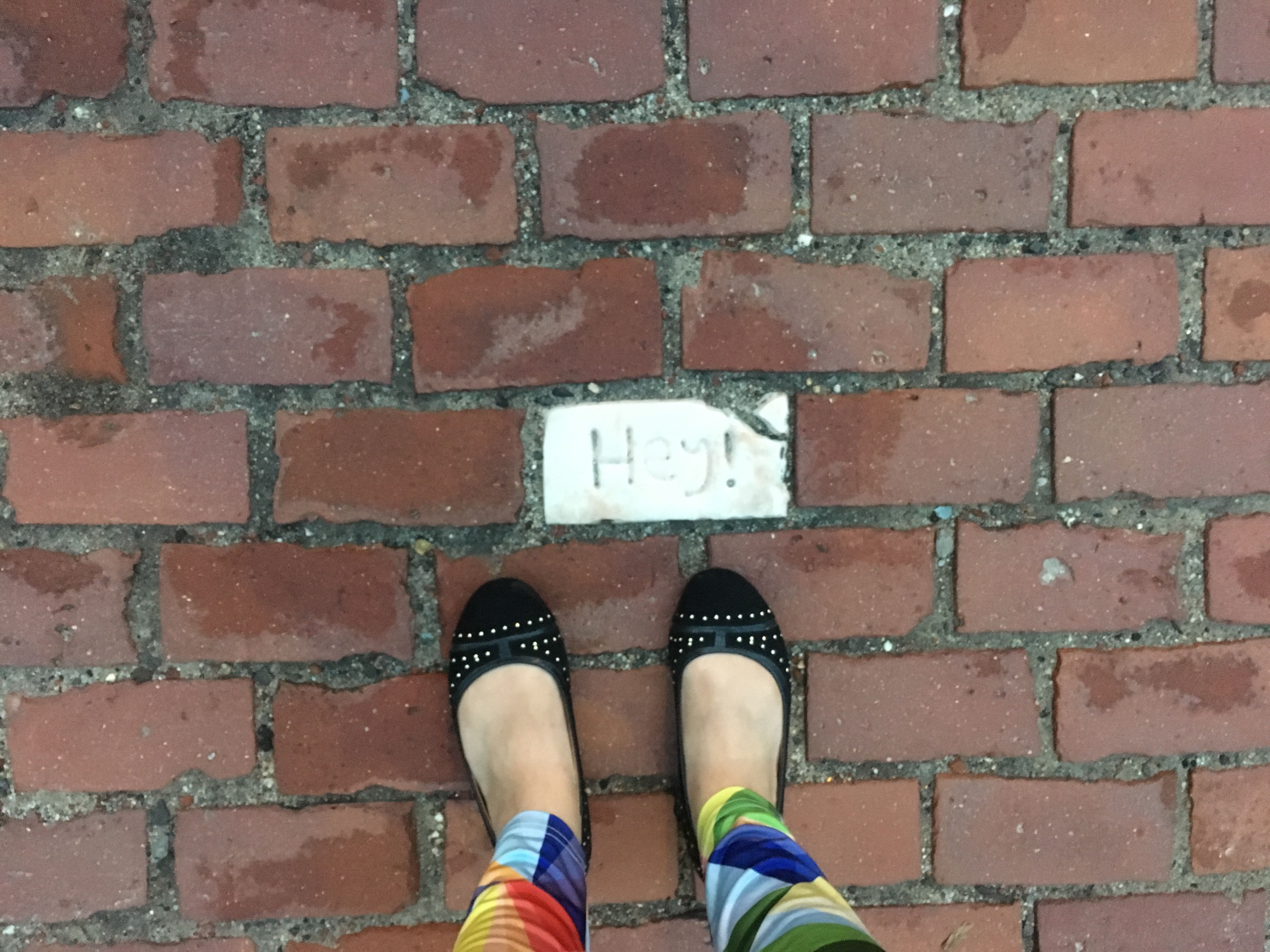 A photograph of feet standing on a brick path. One of the bricks is white and says, "Hey!"