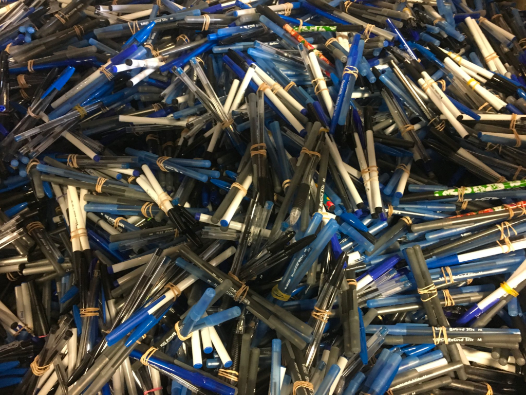 Numerous blue and black pens scattered in a large bin, bundled in groups of three and held together with rubber bands. The entire frame is filled with them so that their numbers overwhlem.